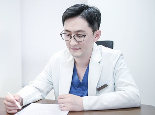 책상에서 서류에 무언가를 쓰는 흰 가운 안경 의사
